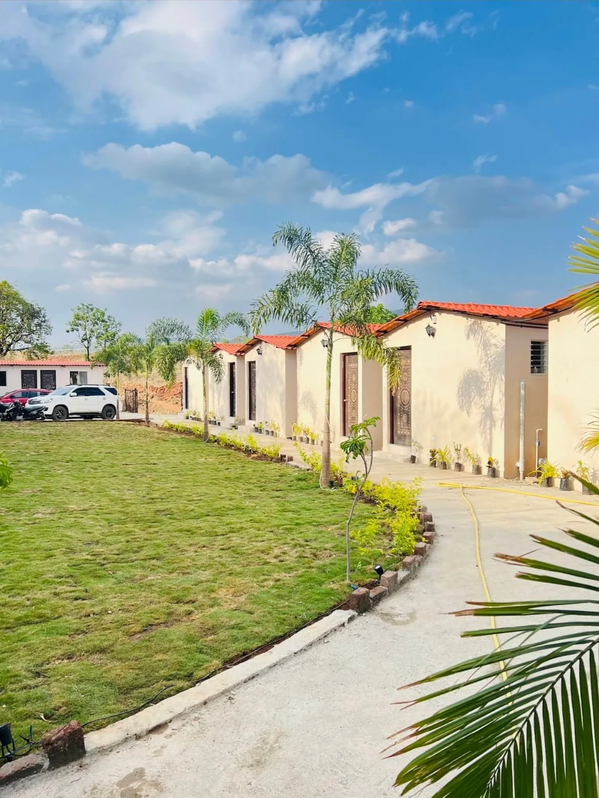 Row of tile-roofed guest cottages with carved doors, facing a landscaped lawn and paved path at Aandra Lakestay Resort