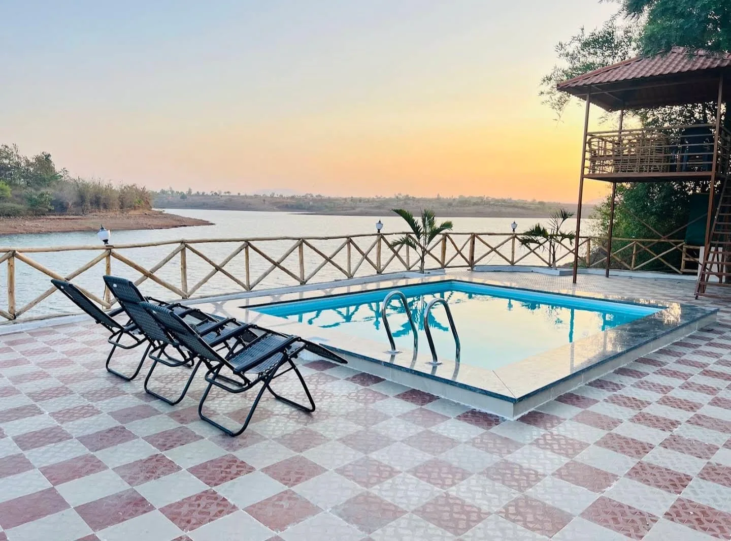 Rectangular swimming pool at the lake edge at sunset, with sun loungers and a bamboo railing, at Aandra Lakestay Resort near Pune, built by PBJ Infra
